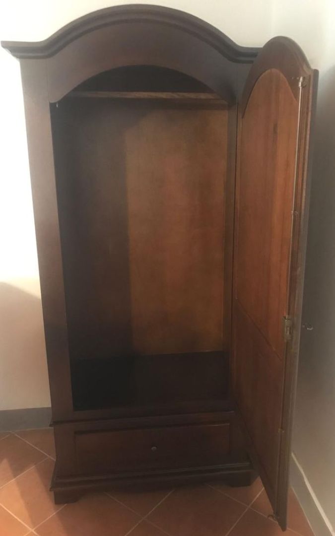 Mobile armadio a un'anta in legno noce scuro con cassetto, posizionato vicino a una finestra.