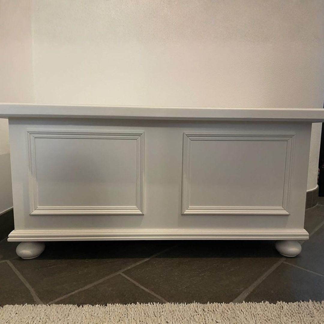 Vista frontale della cassapanca in legno bianco opaco con decorazioni a riquadro e piedi a cipolla classici.