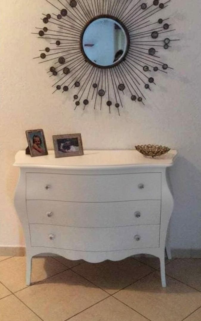 Comò bianco a 3 cassetti in stile classico con linee sinuose, posizionato contro una parete con carta da parati geometrica.