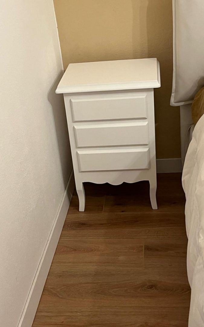 Comodino classico bianco a tre cassetti con gambe sagomate, posizionato accanto a un letto in camera da letto.