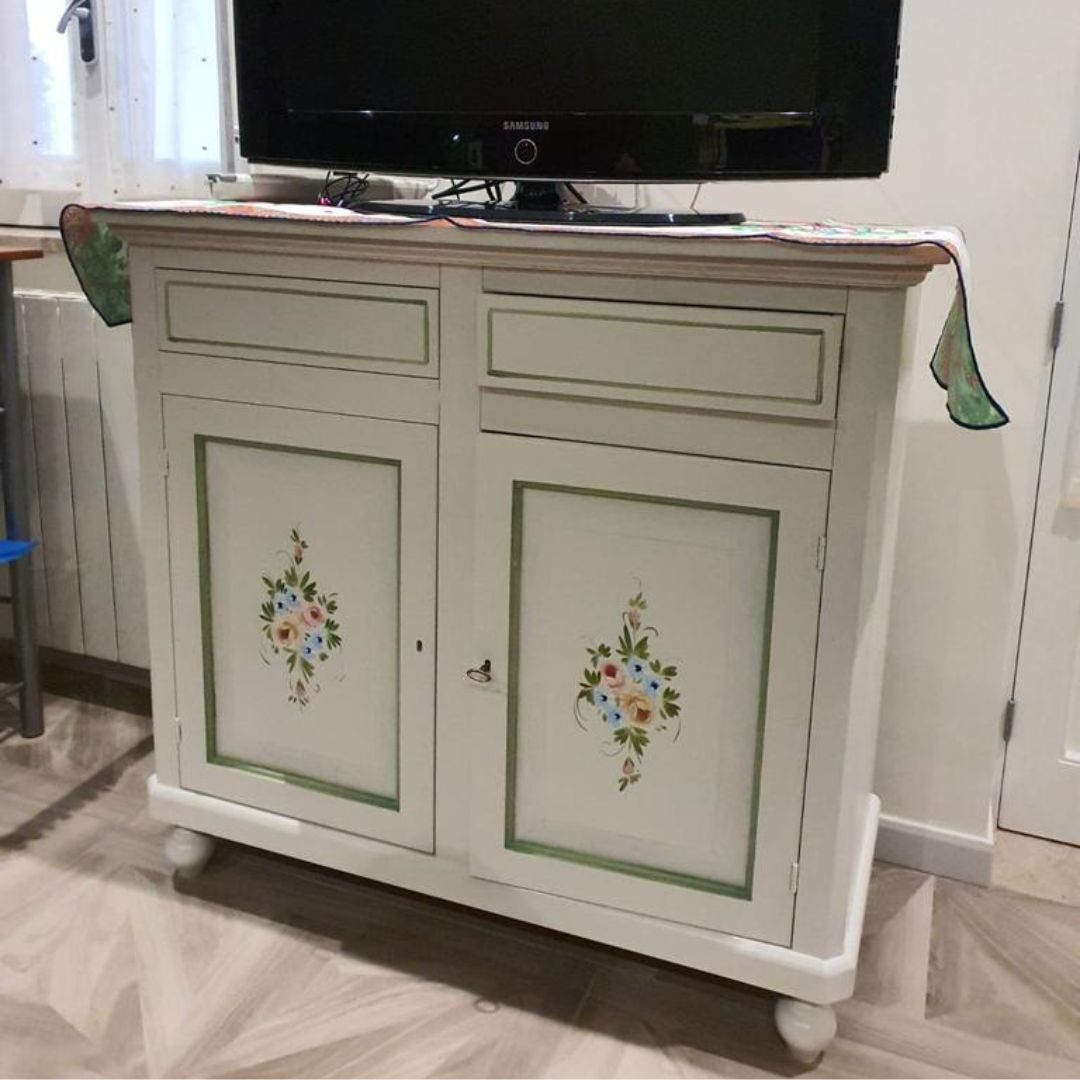 Credenza in legno bianco con due ante, due cassetti e decorazioni floreali dipinte a mano, utilizzata come mobile porta TV.