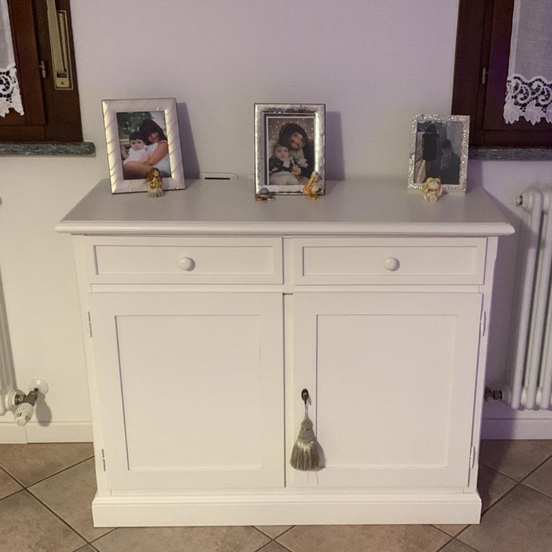 Credenza in legno bianco con due cassetti e ante, decorata con cornici portafoto in un ambiente domestico.