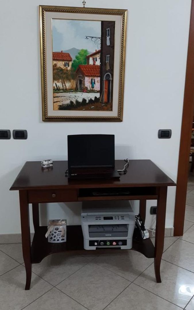 Scrivania in legno scuro con laptop, posta in un ambiente domestico sotto un quadro decorativo.