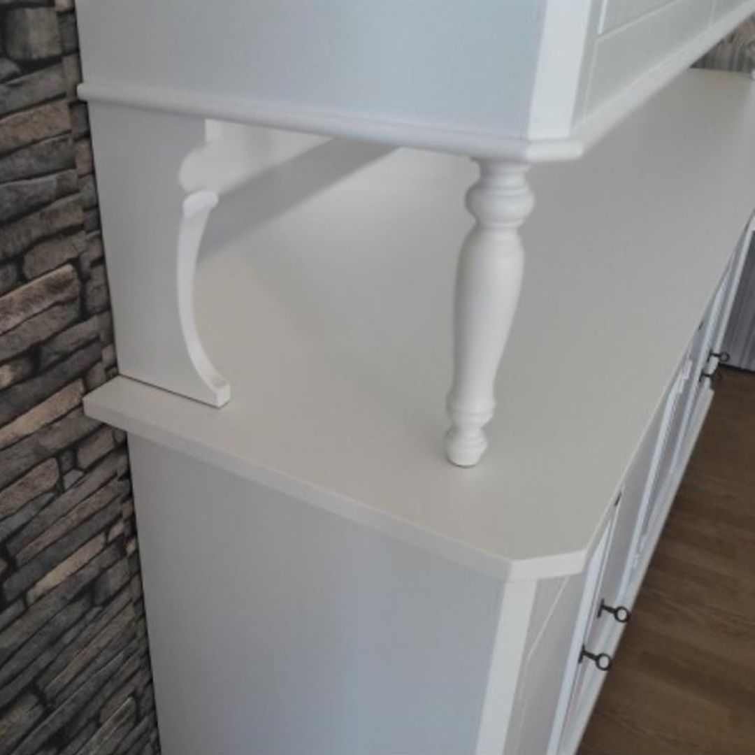 Dettaglio del fianco della vetrina in legno bianco opaco, con vista sul supporto della parte superiore.