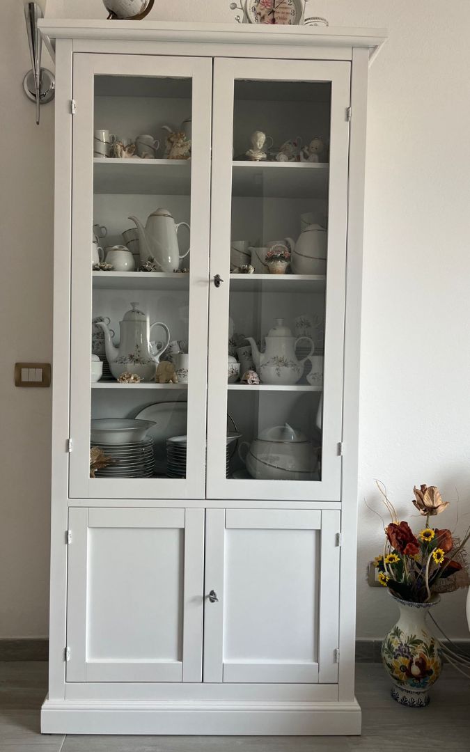 Vetrina classica in legno bianco opaco ricca di decorazioni, bambole e oggetti da collezione.