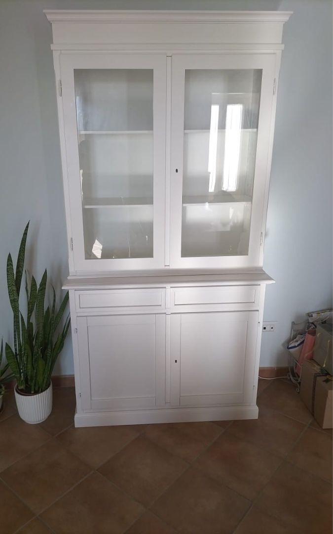 Vetrina in legno bianco posizionata in un soggiorno, con piatti e bicchieri esposti nei vani a giorno.