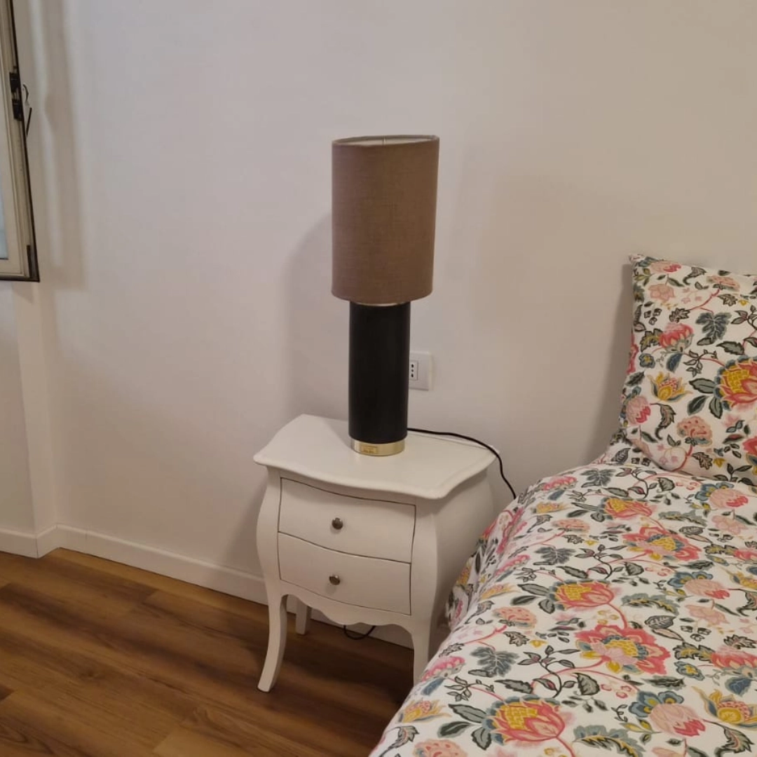 Lampada da tavolo moderna con paralume cilindrico marrone e base nera poggiata su comodino classico bianco in camera da letto - Marca Design Shop