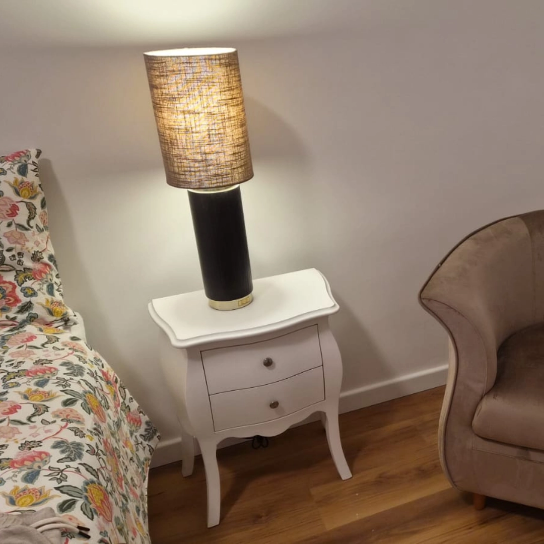 Lampada da comodino accesa con luce calda, paralume in tessuto e struttura cilindrica nera su comodino bianco con cassetti - Marca Design Shop