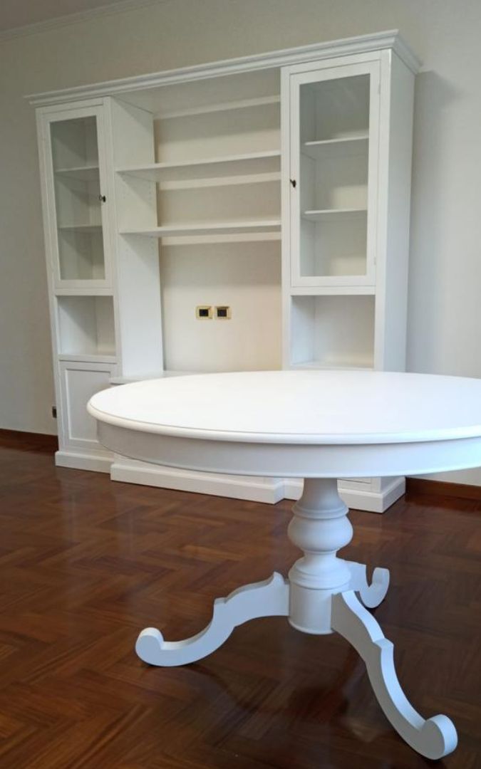 Tavolo rotondo bianco con base lavorata in primo piano e parete attrezzata sullo sfondo.