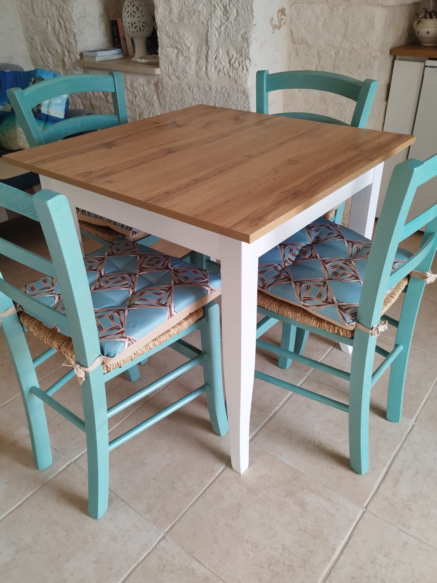 Set tavolo quadrato con piano in legno naturale e struttura bianca, accompagnato da sedie in legno color turchese con cuscini decorati.